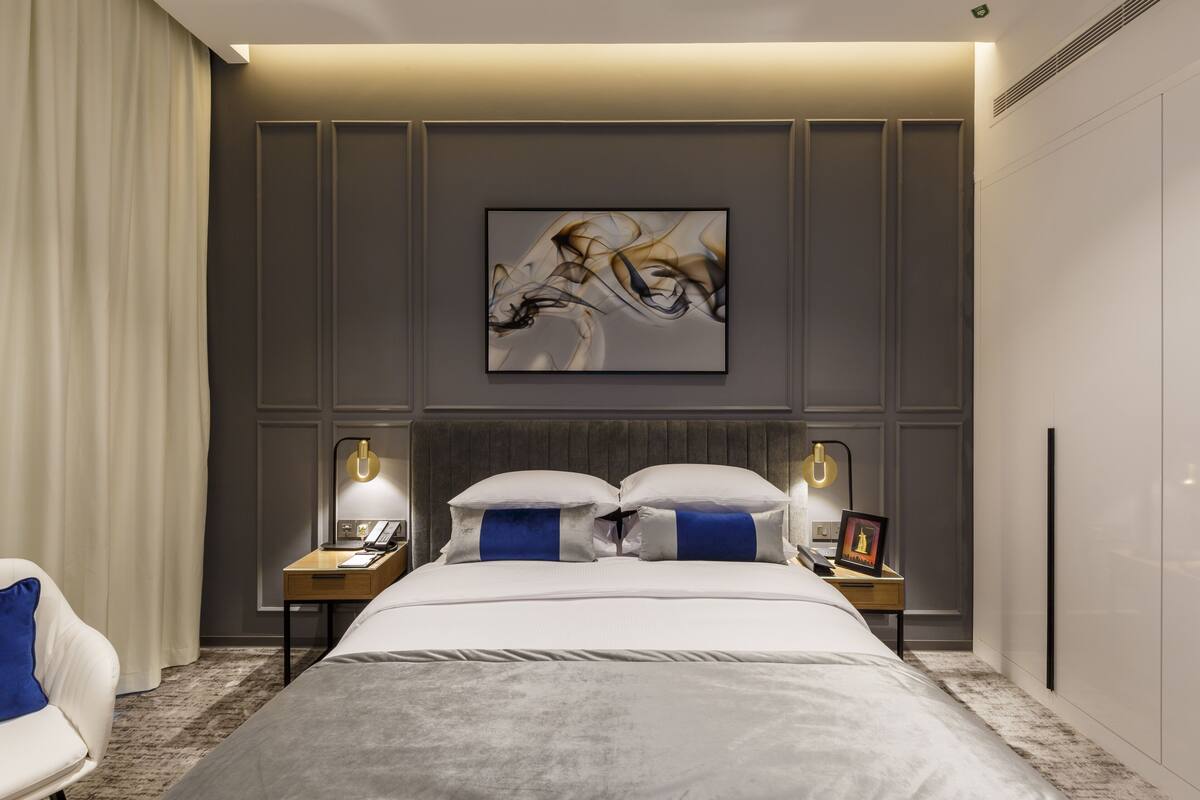 A modern hotel room with a large bed, two bedside tables with lamps, a headboard, and a framed abstract painting on the wall.
