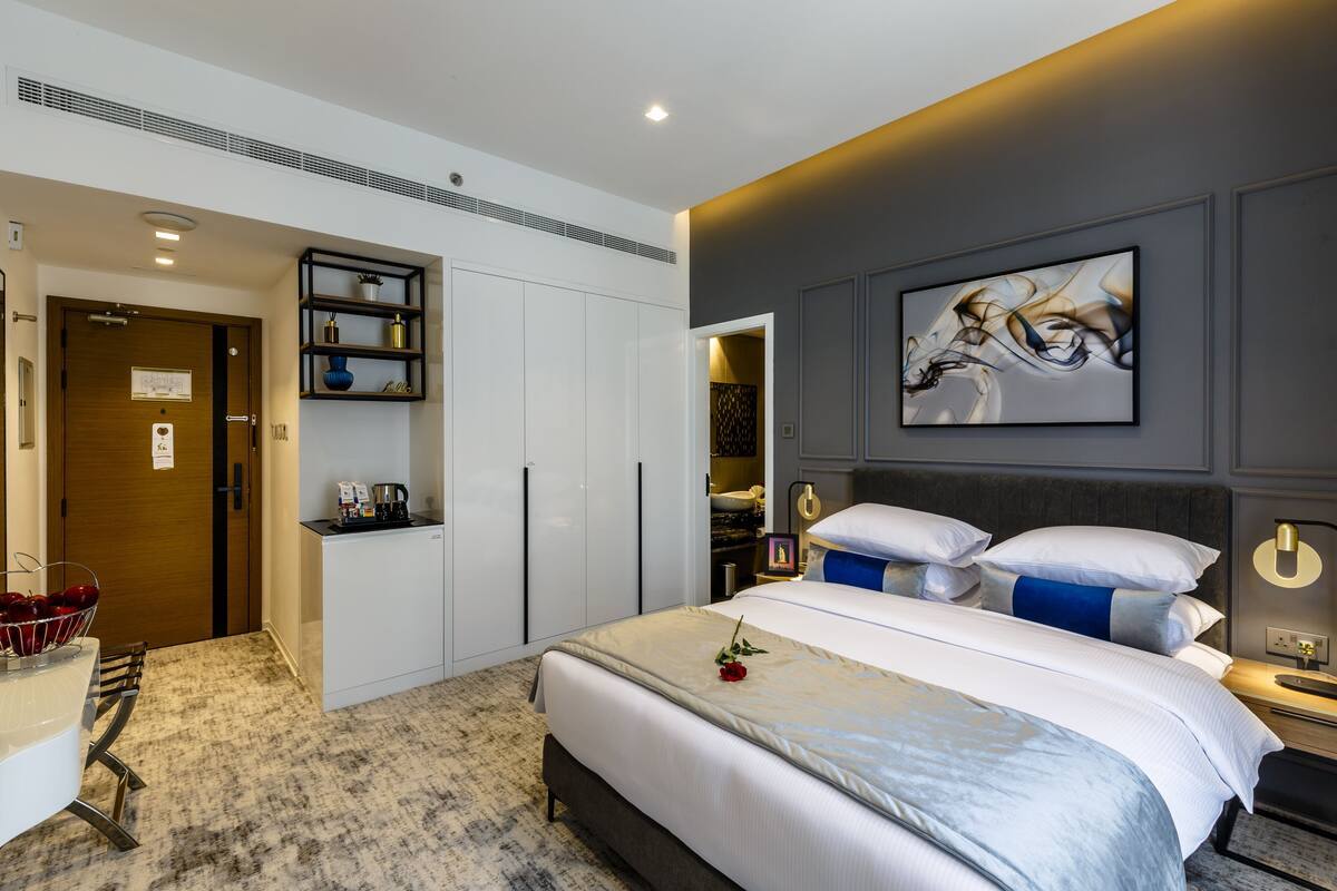 A modern hotel room with a neatly made bed, a small kitchenette, a built-in wardrobe, and a bathroom with a shower and toilet.