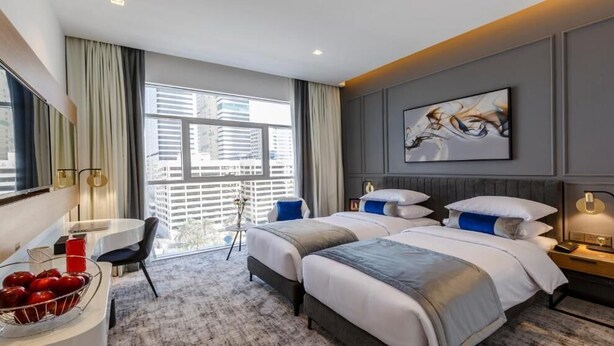 Habitación de hotel con dos camas, un escritorio, una silla, un ventanal con cortinas y un cuadro en la pared.