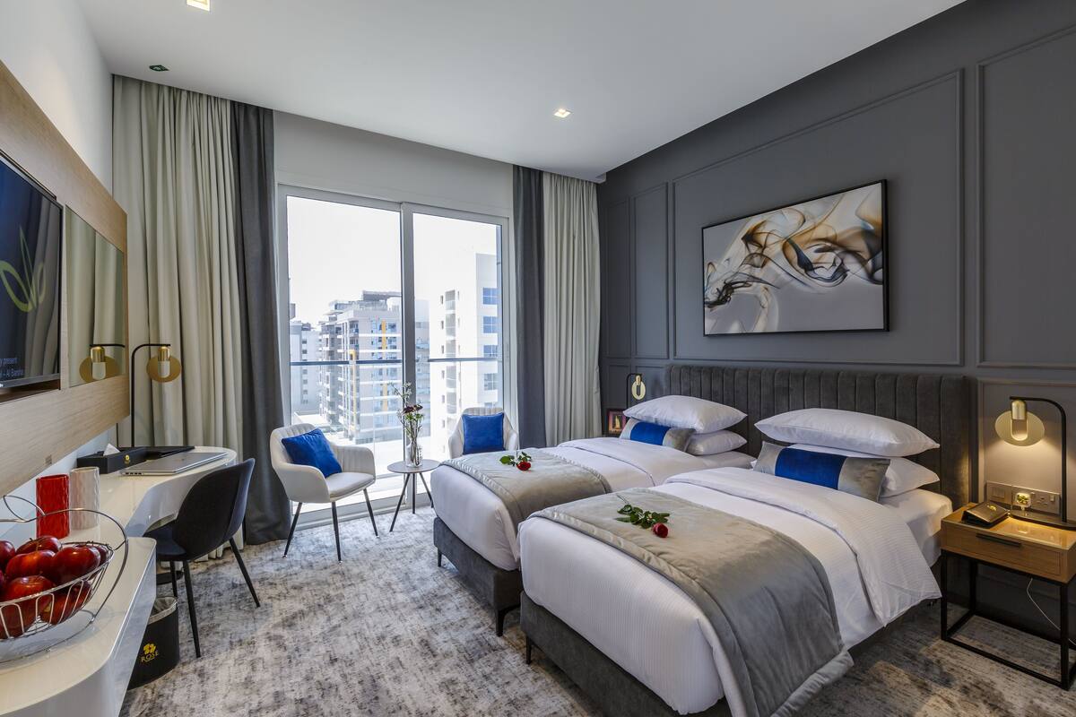 A modern hotel room with two beds, a desk, a chair, a small table with a vase, and a large window offering a city view.