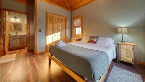 Ein Schlafzimmer mit Holzbalkendecke, einem Bett mit grauer Bettdecke, einem Nachttisch, einem Fenster mit Jalousien und einem Badezimmer mit Spiegel und Waschbecken.
