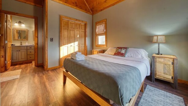 Une chambre avec un plafond en bois, un lit recouvert d’une couverture grise, une table de chevet, une fenêtre avec des stores et une salle de bain avec un miroir et un lavabo.