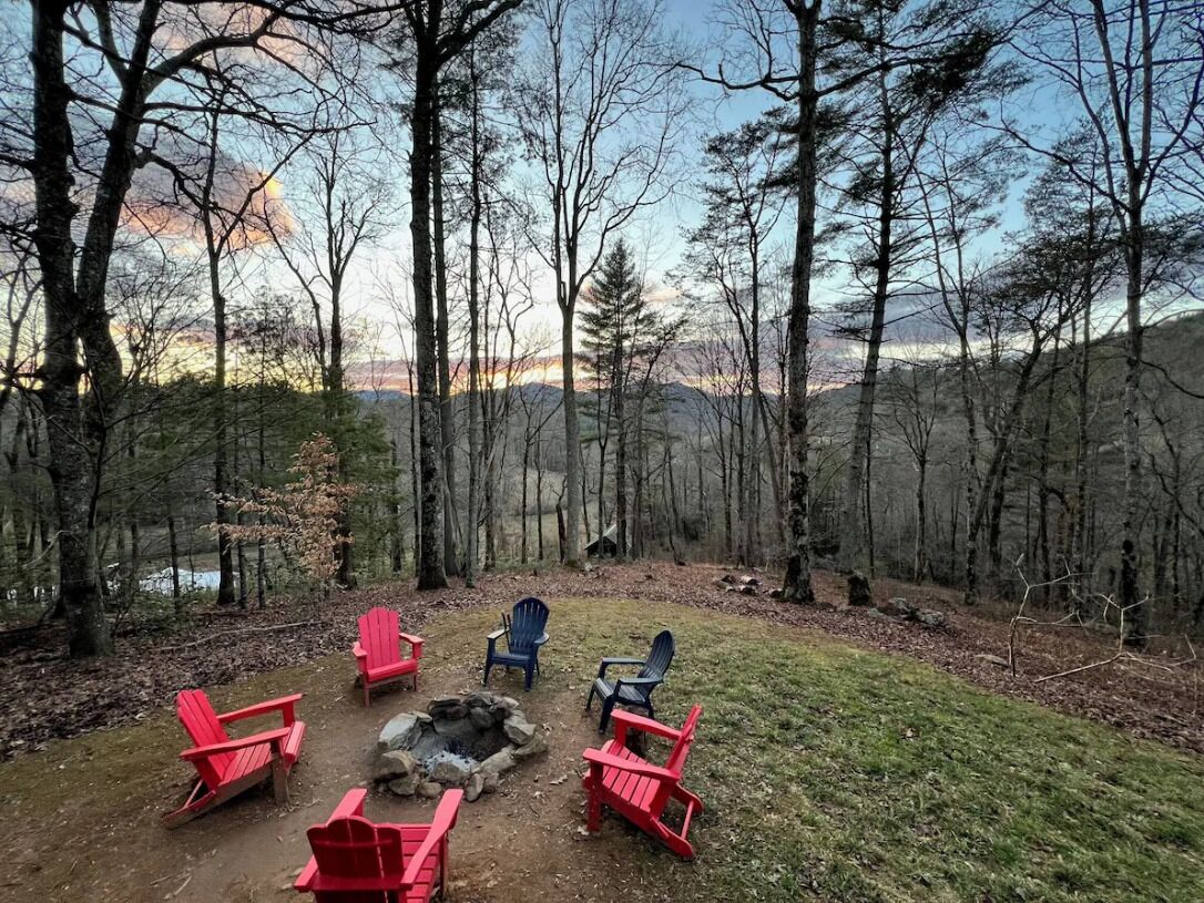Een camping met rode en blauwe Adirondack stoelen, gerangschikt rond een vuurkuil in een bebost gebied, met uitzicht op een meer en bergen op de achtergrond.