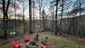 Een camping met rode en blauwe Adirondack stoelen, gerangschikt rond een vuurkuil in een bebost gebied, met uitzicht op een meer en bergen op de achtergrond.