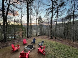 A campsite with red and blue Adirondack chairs arranged around a fire pit in a forested area with a view of a lake and mountains in the background. - Wake to Quiet, Soak Late & Stay a Little Longer (Suches)