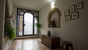 Un couloir avec un sol carrelé, un banc en bois, un petit tabouret, une plante en pot, une étagère fixée au mur avec un vase, et des tableaux encadrés accrochés au mur.