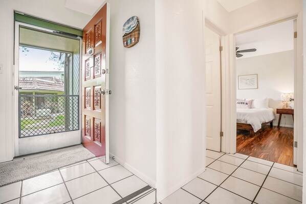 A hallway with a tiled floor, a door with a decorative panel, and a view into a bedroom with a bed and a ceiling fan.