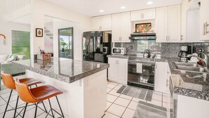 Cocina moderna con gabinetes blancos, encimeras de granito y electrodomésticos de acero inoxidable. Hay dos taburetes de bar y una isla grande.