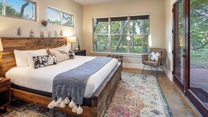 A bedroom with a large bed, wooden headboard, a ceiling fan, and a view of trees through a large window.