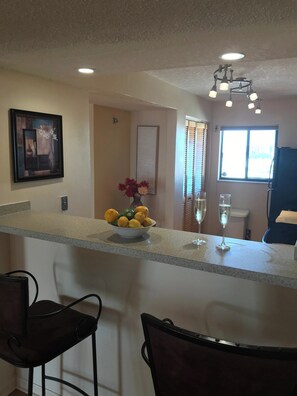 A kitchen with a bar area, a framed artwork, a vase with flowers, and a window with blinds.