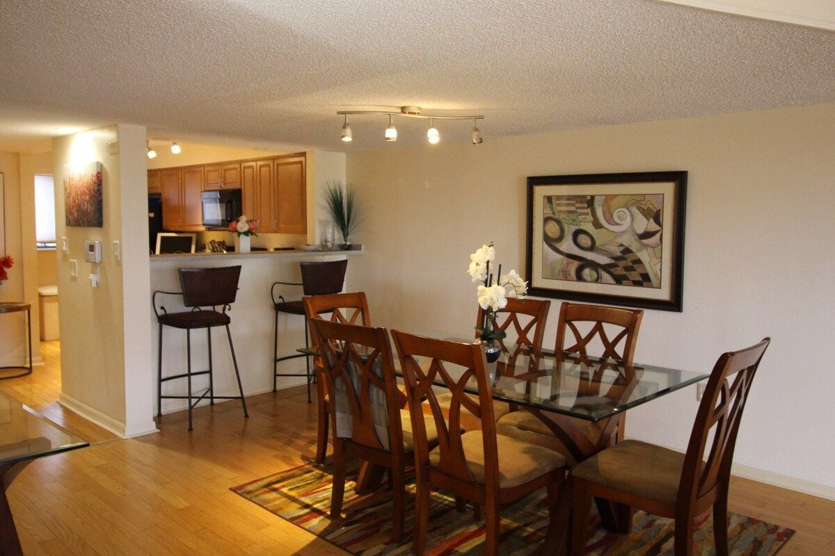 A dining area with a glass table and wooden chairs, a kitchen in the background, and a framed abstract painting on the wall.