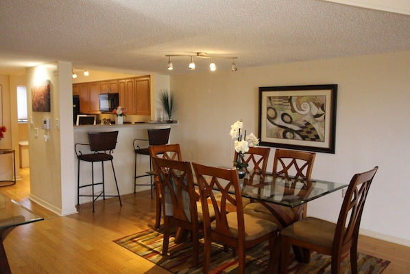 A dining area with a glass table and wooden chairs, a kitchen in the background, and a framed abstract painting on the wall.