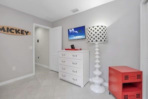 A hallway with a white dresser, a red storage unit, a Mickey-themed sign, a TV on the wall, and a polka-dotted lamp.