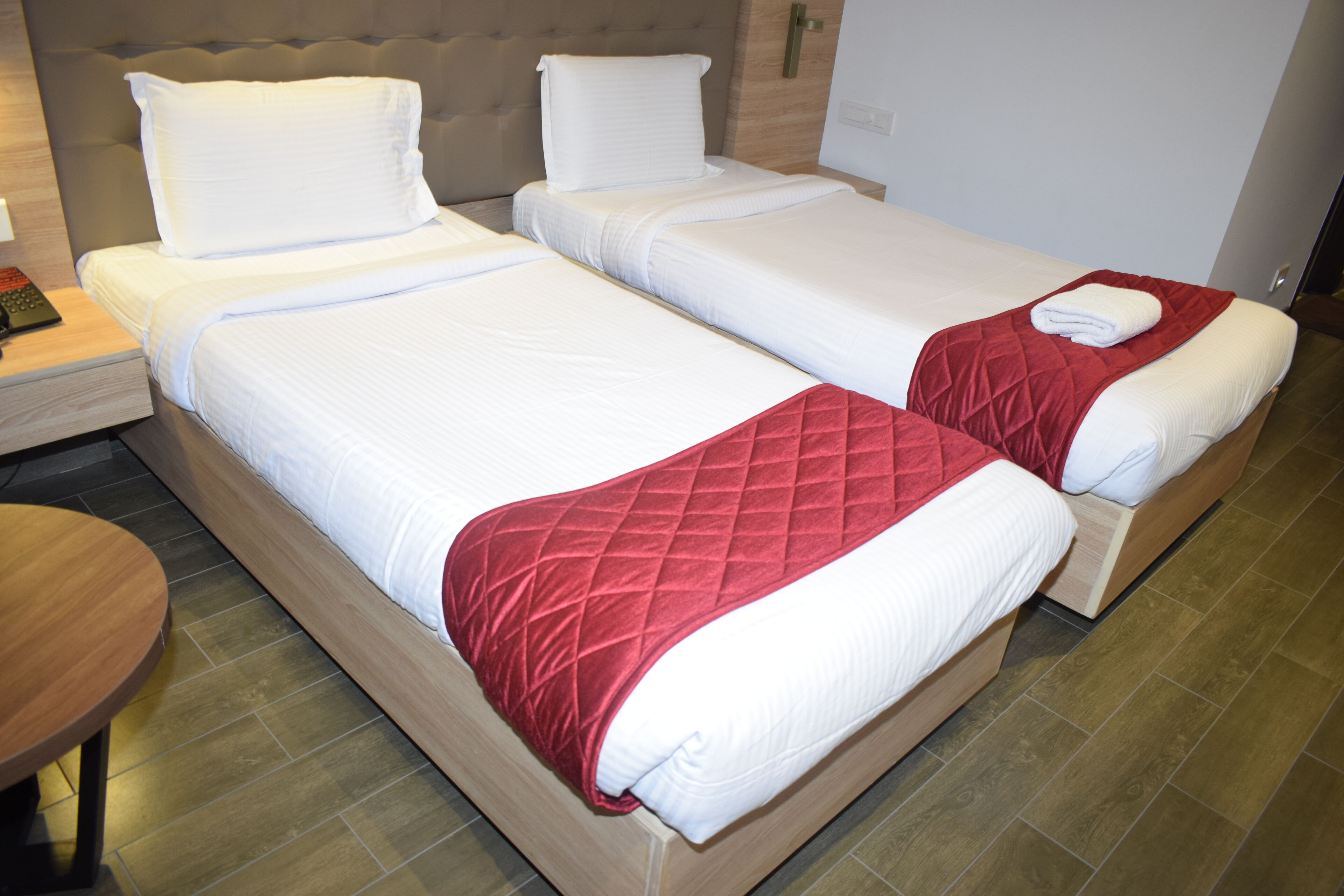 A hotel room with two single beds, each with white bedding and a red quilted throw. There is a wooden nightstand with a remote control on it, and a round wooden table. The room has a modern design with a grey upholstered headboard and a wooden floor.