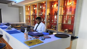 A bar area with a counter, shelves with bottles, and a person working behind the bar. - Hotel Annamalai (Bahur)