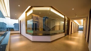 A modern hotel corridor with a glass-walled corner office, a staircase, and a long hallway with doors on one side. - Hotel Annamalai (Bahur)