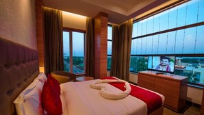 A hotel room with a large bed, a TV on a wooden dresser, and a view of the city through large windows. - Hotel Annamalai (Bahur)