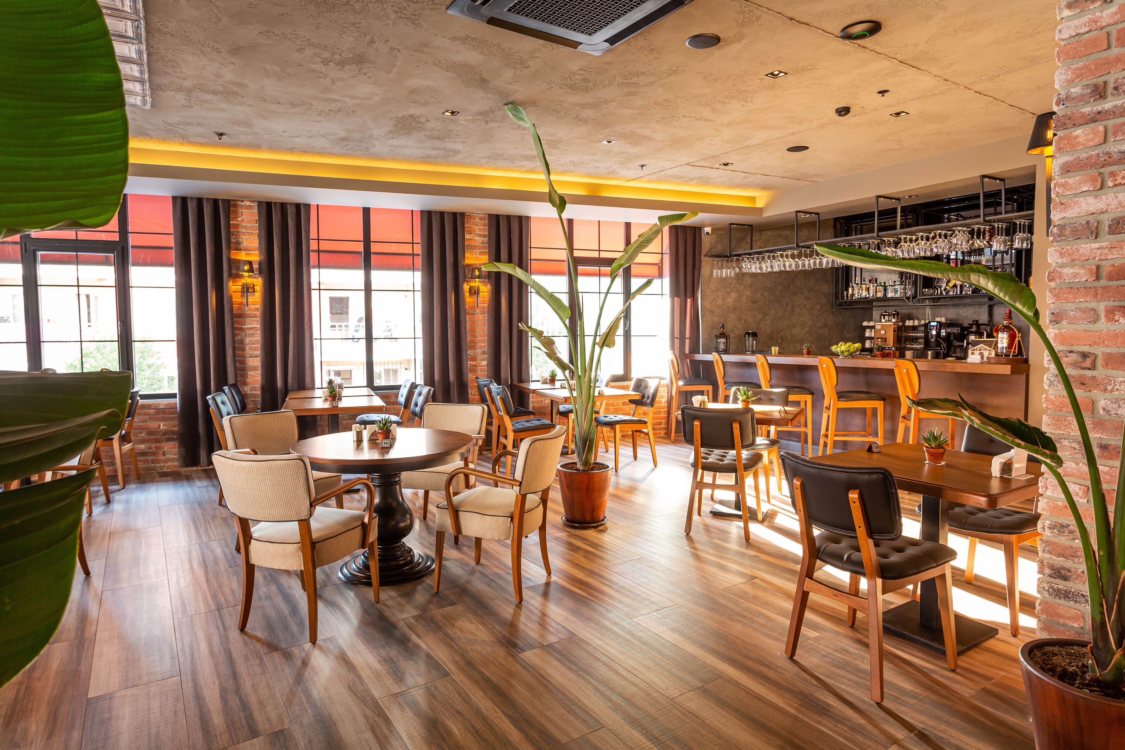 A modern restaurant with wooden flooring, multiple dining tables, and chairs. There are potted plants and a bar area with shelves of bottles.