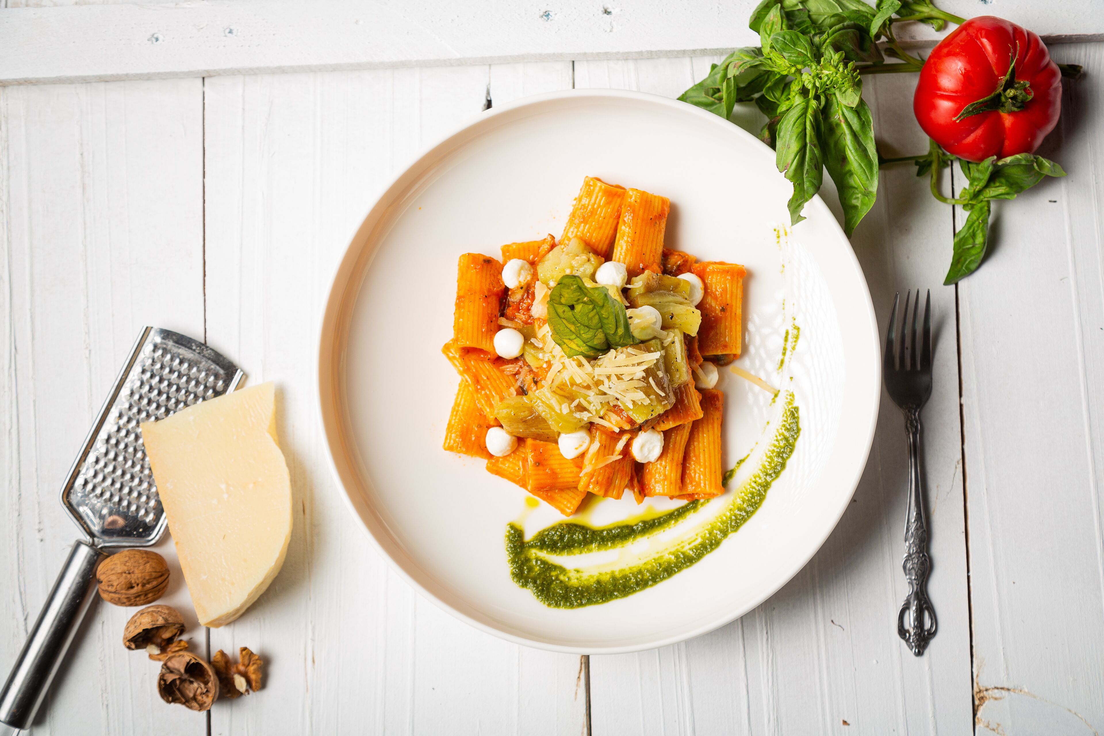 A plate of pasta with cheese and nuts, a grater, walnuts, and a tomato on a white wooden table.