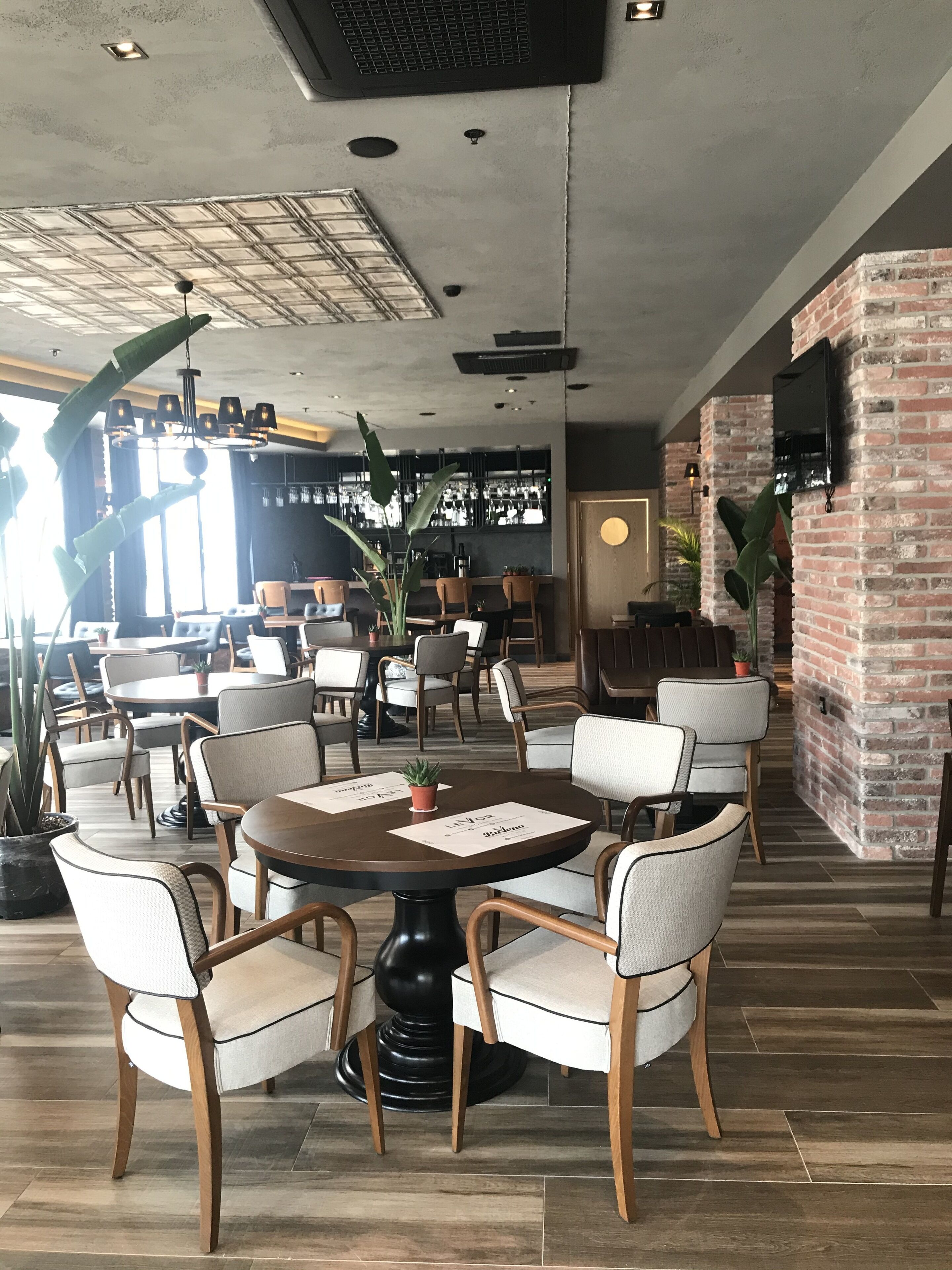 A restaurant interior with round tables, cushioned chairs, a brick accent wall, and a bar area in the background.