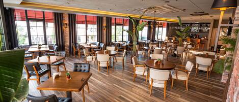 Un restaurante espacioso con varias zonas de comedor, mesas y sillas de madera, y amplios ventanales que dejan entrar luz natural.