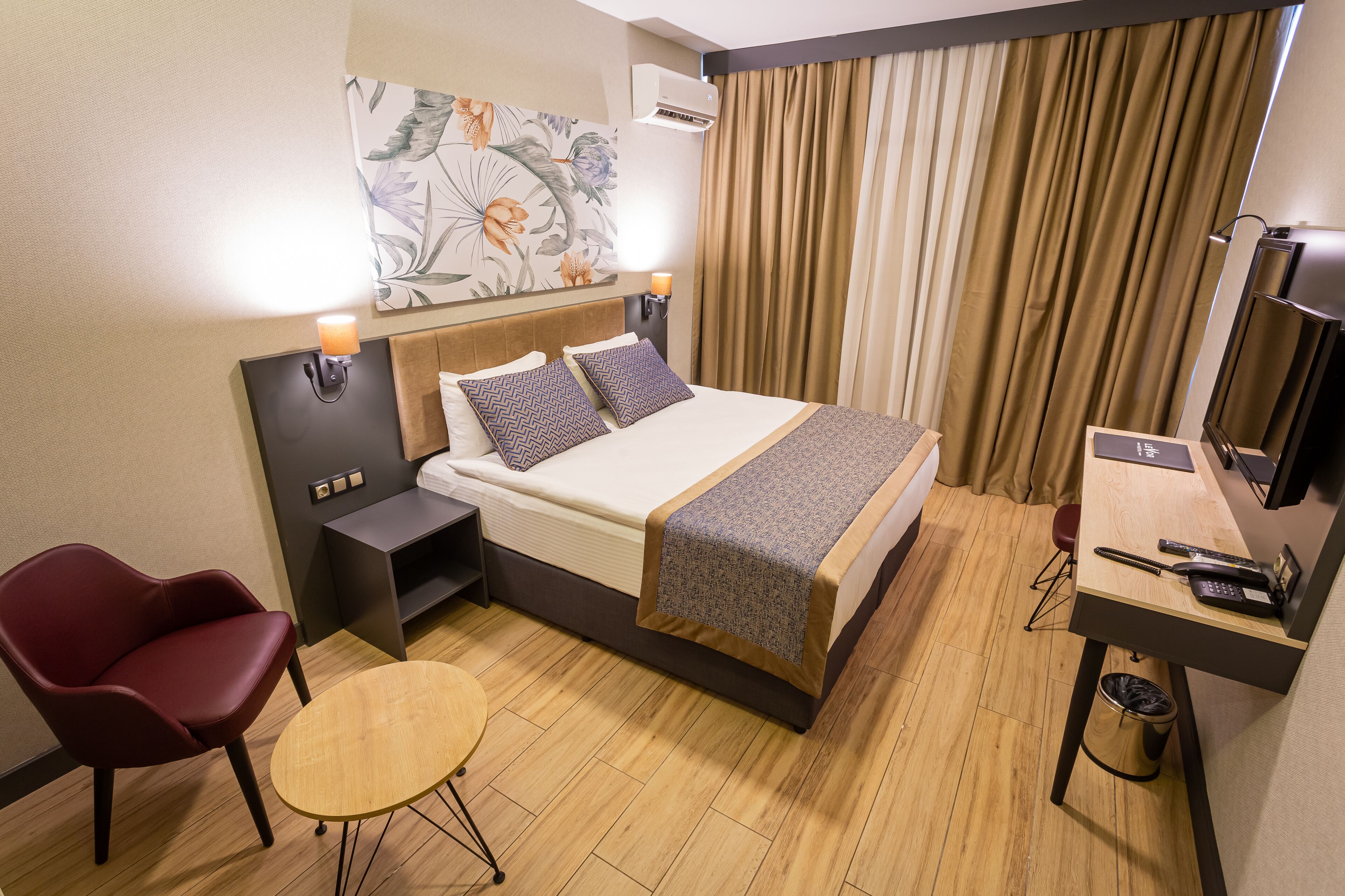 A hotel room with a bed, a desk, a chair, a small table, and a painting on the wall.
