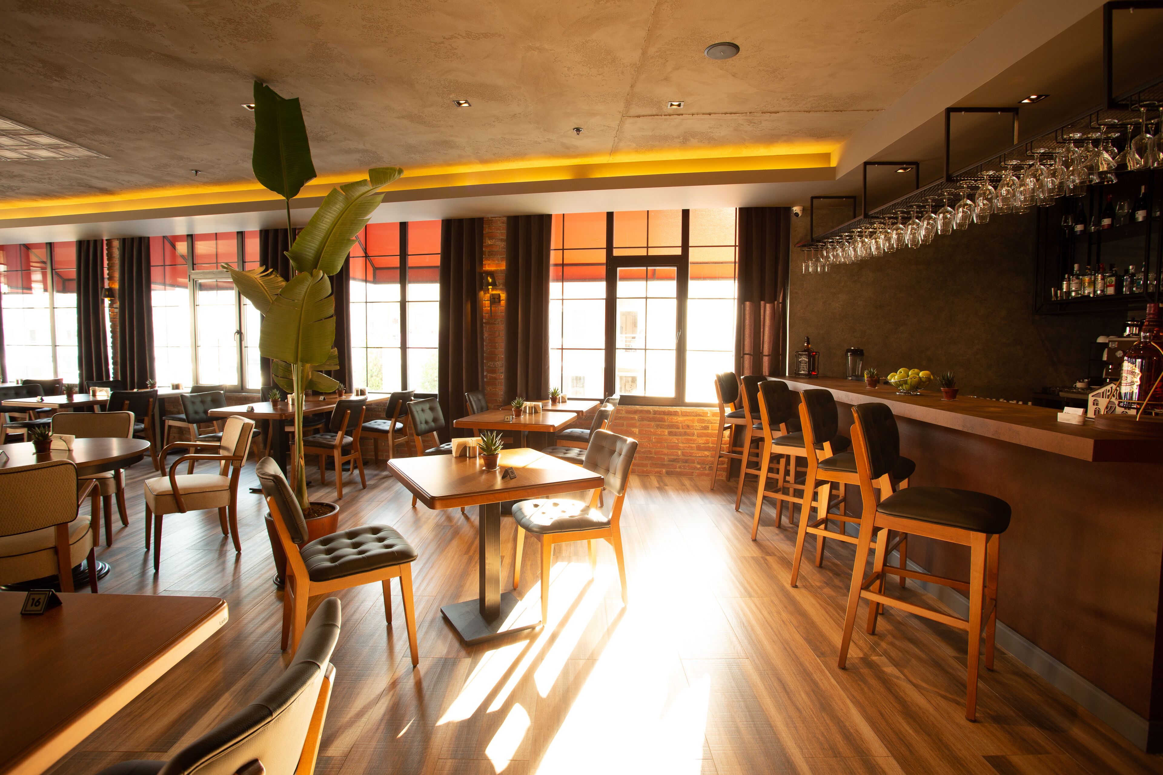 A restaurant with wooden tables and chairs, a bar area with glasses, and large windows allowing natural light.