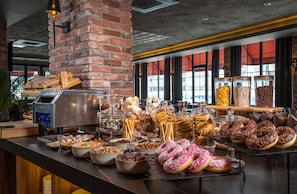A buffet with various food items, including donuts and pastries, arranged on a counter with a brick wall and large windows in the background. - Levor Hotel (Bursa)