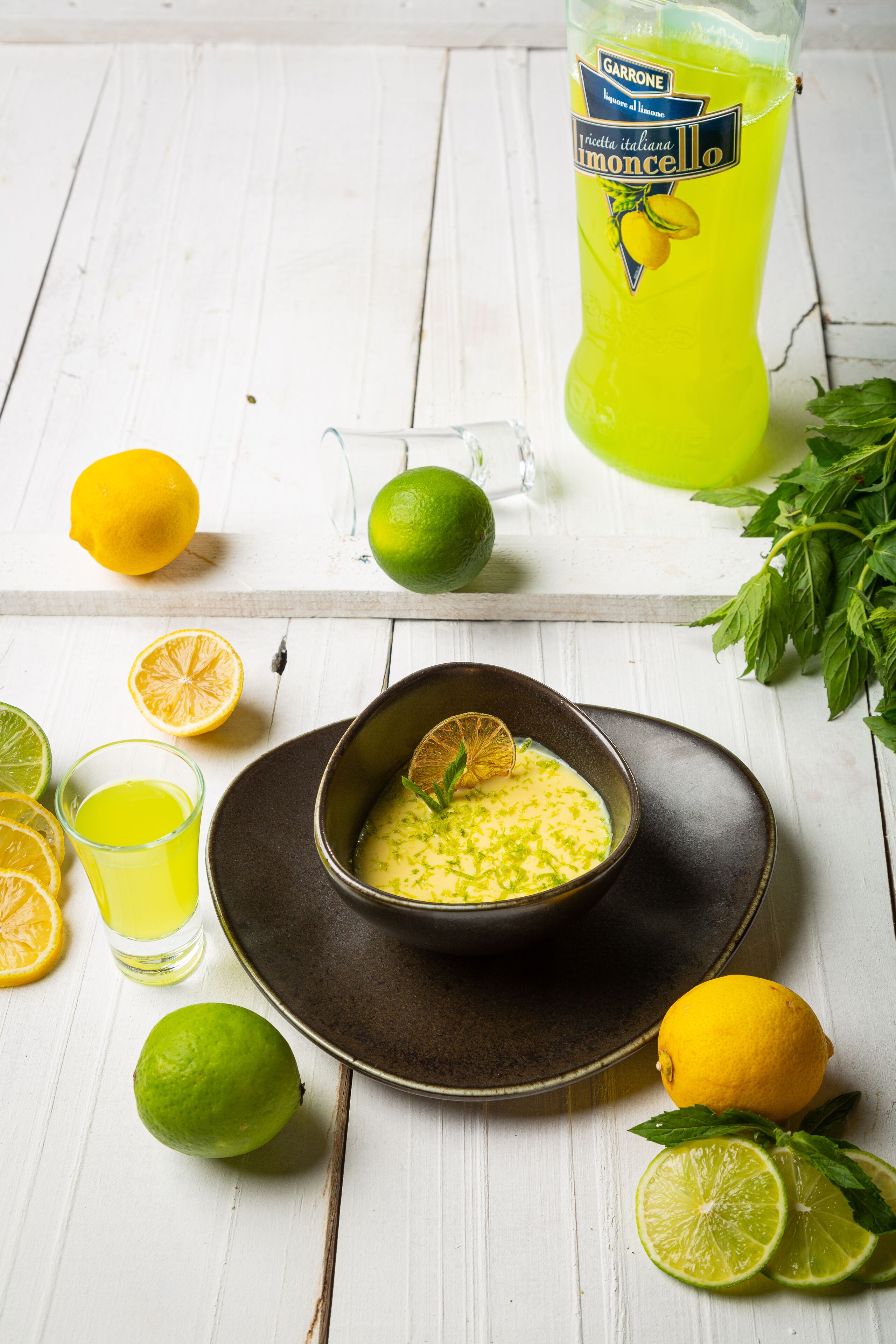 A bowl of yellow soup with a lemon garnish, surrounded by limes and a glass of yellow liquid on a white wooden table.