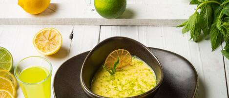 A bowl of yellow soup with a lemon garnish, surrounded by limes and a glass of yellow liquid on a white wooden table.