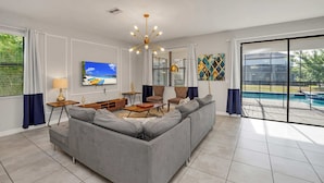 A spacious living room with a grey sofa, a coffee table, and a TV mounted on the wall. There is a large window with a view of a swimming pool and greenery outside. - Grand, Luxurious Home, Toy Story Room and Big Pool Area #9cg476 (Davenport)
