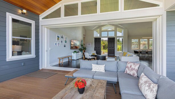 A spacious living area with a grey sofa, wooden coffee table with flowers, and a view of the backyard through large windows. - Nakoa Kai in Sunny Poipu with Pool (Koloa)