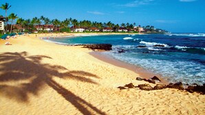 A beach with clear blue water, palm trees, and a row of buildings along the shore. - Nakoa Kai in Sunny Poipu with Pool (Koloa)