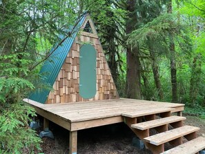 Una estructura de madera tipo cabaña con techo de lona verde y plataforma plana, rodeada de un bosque denso.