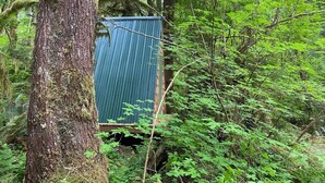 Pieni, sininen, aaltopellistä rakennettu rakennelma sijaitsee metsäisellä alueella, ja sen ympärillä on tiheää, vihreää kasvillisuutta.