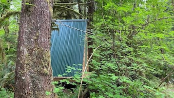 A small, blue, corrugated metal structure in a forested area, surrounded by dense green foliage.