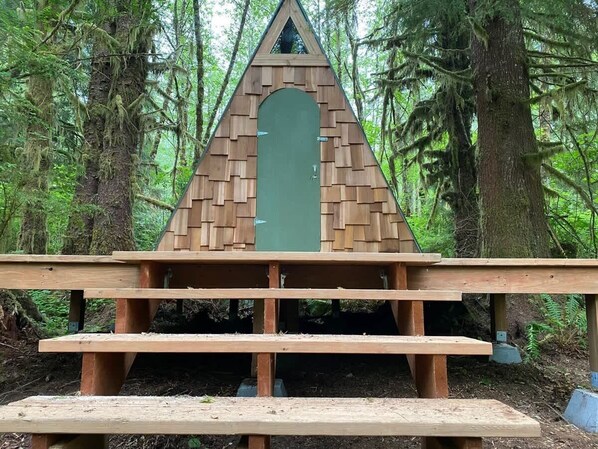 A wooden structure with a triangular roof and a green door, surrounded by a forest.
