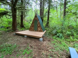 Een klein houten vakantiehuisje met een schuin dak en een groene deur, gelegen in een bebost gebied met bomen en begroeiing.