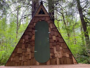 A triangular wooden structure with a green door, surrounded by a forest.