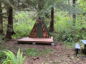 Een houten constructie in een bos, mogelijk een prieel of schuilplaats, met een groen dak en een driehoekig ontwerp.