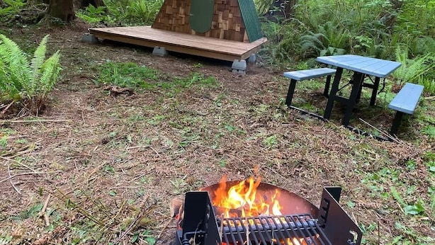 Een bebost gebied met een klein houten gebouw, een vuurplaats en een picknicktafel.