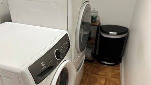 A compact laundry area with a front-loading washer and dryer, a trash bin, and a small shelf with items.