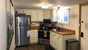 A compact kitchen with white cabinets, stainless steel appliances, a wooden countertop, and a black trash bin.