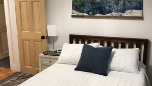 A neatly made bed with a blue blanket and two pillows, a bedside lamp, and a painting of a mountain landscape on the wall.