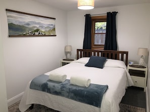 Cama bem arrumada com duas toalhas enroladas, pintura de montanhas na parede, janela com cortinas e luminárias de cabeceira.