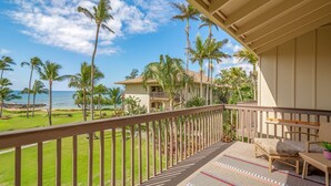 A balcony with a view of the ocean and palm trees, a lounge area with a sofa and a small table, and a building with a balcony in the background. - Lae Nani 434 1 Bedroom Condo by RedAwning (Kapaa)