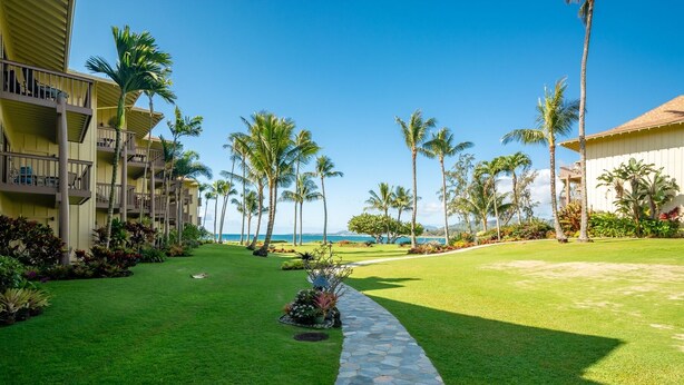 A hotel courtyard with a stone pathway leading to a pool, surrounded by palm trees and a view of the ocean. - Lae Nani 434 1 Bedroom Condo by RedAwning (Kapaa)
