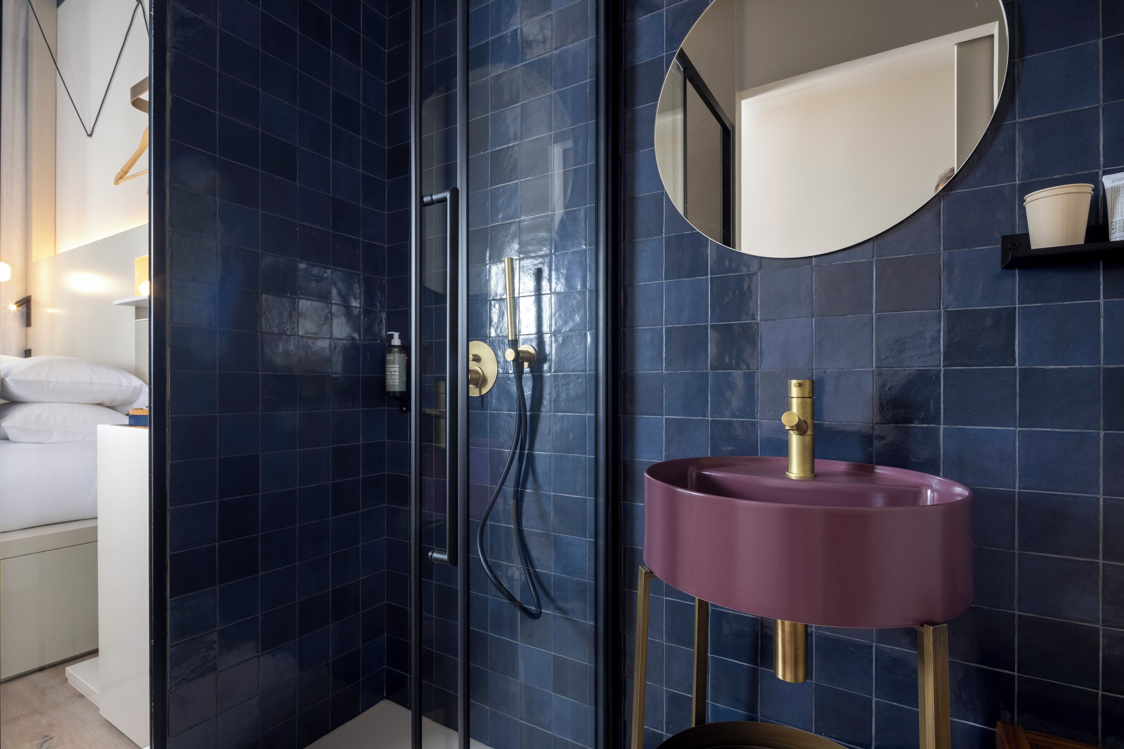 Une salle de bain moderne avec des carreaux bleu foncé, une douche en verre, un lavabo rose agrémenté de robinetterie dorée et un miroir rond accroché au mur.