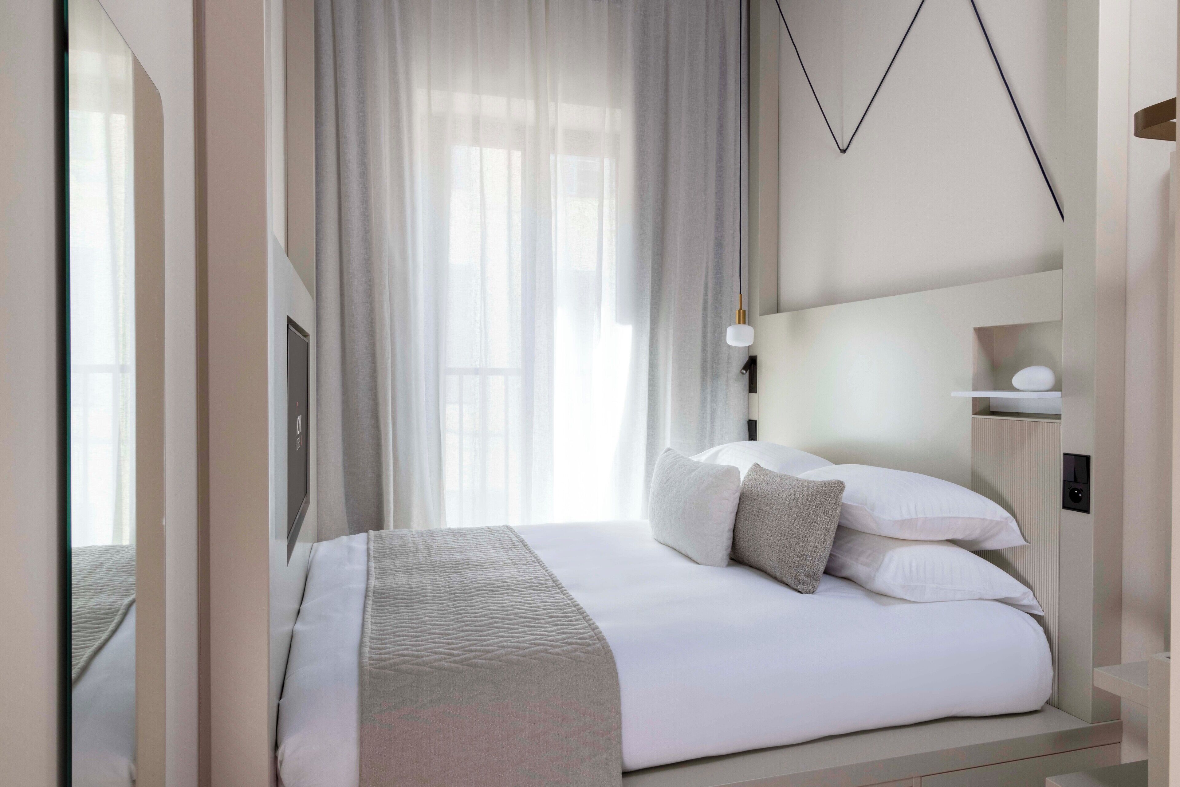 A modern hotel room with a neatly made bed, a large window with sheer curtains, and a minimalist design.