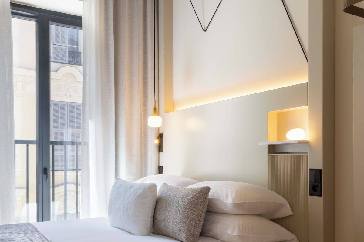 Ein Hotelzimmer mit Bett, Nachttisch, Lampe und Blick auf ein Gebäude durch ein Fenster.